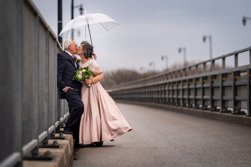 Couple d’âge mûr qui s’embrasse sous un parapluie transparent sur un pont, photographe renouvellement de vœux
